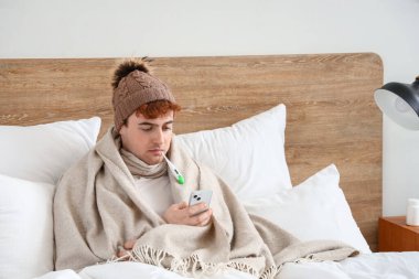 Yatak odasında cep telefonu kullanan termometresi olan hasta bir genç.