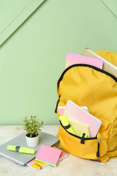 靠近绿墙的浅绿色桌子上有带笔记本 家居用品和标识的黄色校服背包 — 图库照片