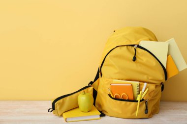 Sarı okul çantası, duvarın yanındaki tahta masada defter, elma ve makas.