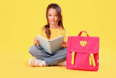 Sarı arka planda kitap ve sırt çantası olan küçük okul kızı.