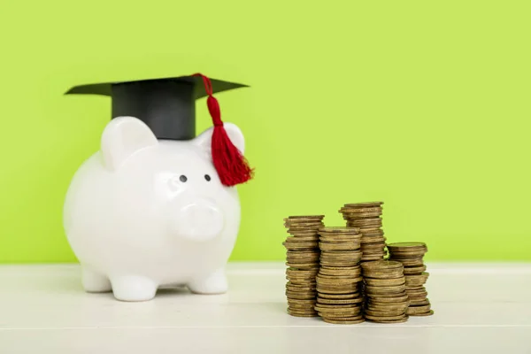 卒業キャップと緑の背景に白いテーブルの上にコインのスタックを持つ貯金箱 学生ローンの概念 — ストック写真