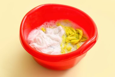 Kirli elbiseli plastik leğen, sarı arka planda su ve köpük.