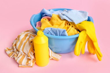 Kirli çamaşırlı plastik leğen, bir şişe çamaşır deterjanı ve pembe arka planda sarı lastik eldivenler.