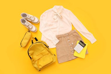 Şık okul üniforması, sırt çantası, spor ayakkabısı ve turuncu arka planda kırtasiye malzemesi.