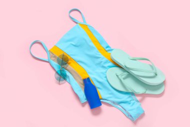 Şık bir mayo, parmak arası terlik, güneş gözlüğü ve pembe arka planda bir şişe güneş kremi.