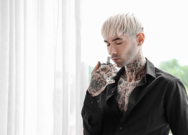 Evinin penceresinin yanında parfümlü dövmeli bir genç.