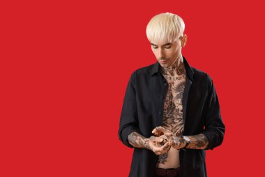 Dövmeli genç adam kırmızı arka plana parfüm sıkıyor.