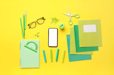 Gözlüklü cep telefonu ve sarı arka planda farklı kırtasiye malzemeleri.