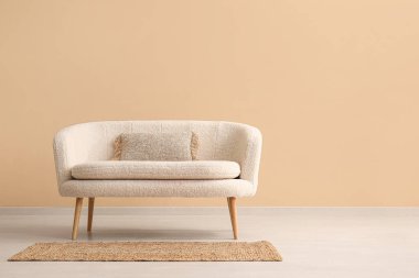 Bej duvarın yanında yastıklı ve halılı rahat beyaz kanepe.