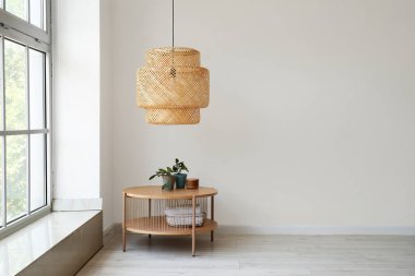 Ev bitkisi ve hasır lambalı ahşap sehpa beyaz duvarın yanında.