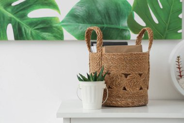 Beyaz duvarın yanındaki çekmecelerin üzerinde kitap ve çiçek sepeti var.