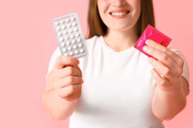 Pembe arka planda prezervatifli ve doğum kontrol haplı genç kadın, yakın plan.