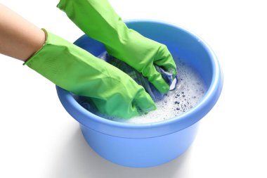 Yeşil lastik eldivenli bir kadın beyaz arka planda plastik bir leğende çamaşır yıkıyor.
