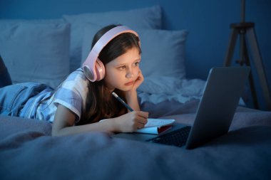 Kulaklıklı küçük kız geceleri yatak odasında internetten ders çalışıyor.