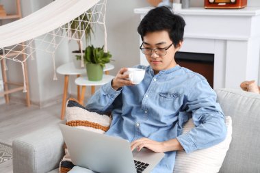 Evde boş gününde dizüstü bilgisayar kullanan Asyalı genç bir adam.