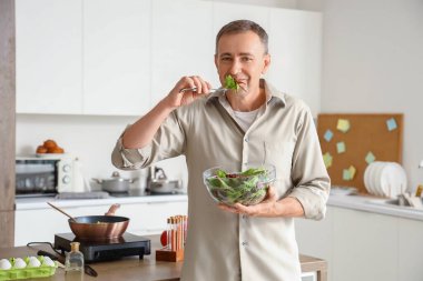 Olgun bir adam mutfakta sebze salatası yiyor.