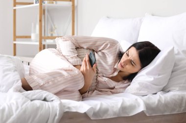 Cep telefonlu genç bir kadın yatak odasında regl sancısı çekiyor.