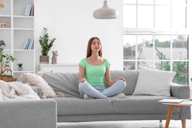 Oturma odasının içindeki gri kanepede meditasyon yapan güzel bir kadın.
