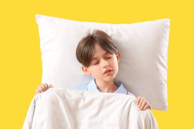 Sarı arka planda yastıklı ve battaniyeli uyuyan küçük çocuk.