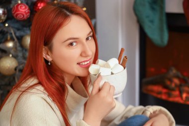 Noel arifesinde şöminenin yanında kakao içen genç bir kadın.