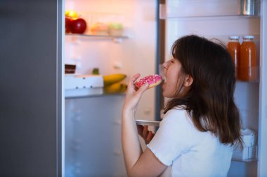 Genç bir kadın geceleri açık buzdolabının yanında lezzetli çörekler yiyor.