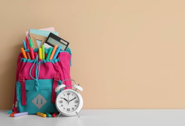 Turuncu duvarın yanındaki beyaz masanın üzerinde farklı kırtasiye malzemesi ve alarm saati olan renkli okul çantası.