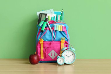 Yeşil duvarın yanındaki ahşap masada farklı kırtasiye malzemesi ve alarm saatleriyle renkli okul çantası.