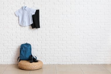 Sırt çantalı, ayakkabılı ve şık okul üniformalı puf. Odadaki açık tuğlalı duvarda asılı.