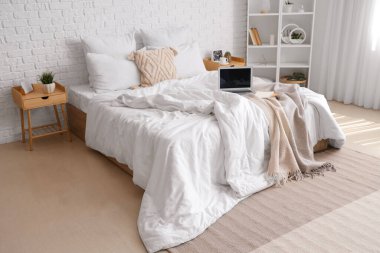 Beyaz battaniyeli dağınık yatak, defterler ve aydınlık yatak odasında modern dizüstü bilgisayar.