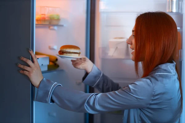 Mladá Žena Chutným Hamburgerem Blízkosti Otevřené Lednice Kuchyni Noci — Stock fotografie