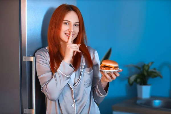Młoda Kobieta Smacznym Hamburgerem Pokazuje Gest Milczenia Pobliżu Otwartej Lodówki — Zdjęcie stockowe