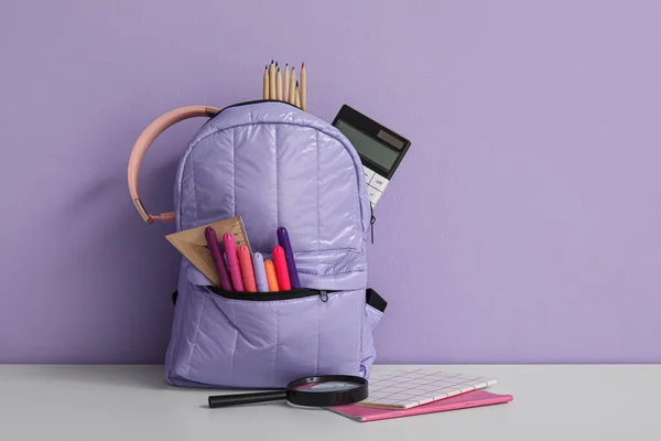 紫色墙壁附近白色桌子上装有不同文具和耳机的学校背包 — 图库照片
