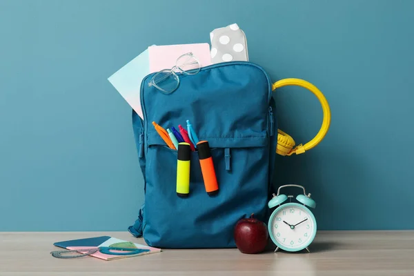 青い壁の近くの木製テーブルの上に異なる文房具や目覚まし時計とカラフルな学校のバックパック — ストック写真
