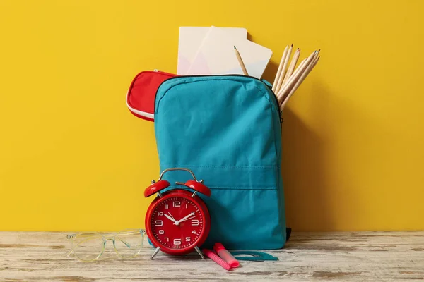 蓝色校服背包 装有不同的文具和黄色墙壁附近木制桌子上的闹钟 — 图库照片