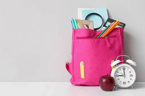 粉红校服背包 装有不同的文具 闹钟和新鲜苹果 放在灰墙附近的白桌子上 — 图库照片
