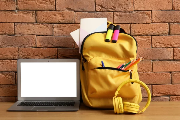 Yellow school backpack with stationery, laptop and headphones on wooden table near brown brick wall