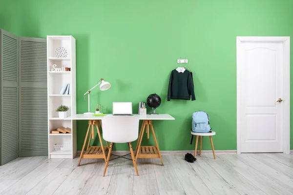 stock image Workplace with desk, shelving unit, laptop computer, backpack and stylish school uniform hanging on green wall