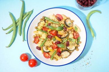 Bir kase lezzetli makarna salatası, domatesli ve mavi arka planda fesleğen.