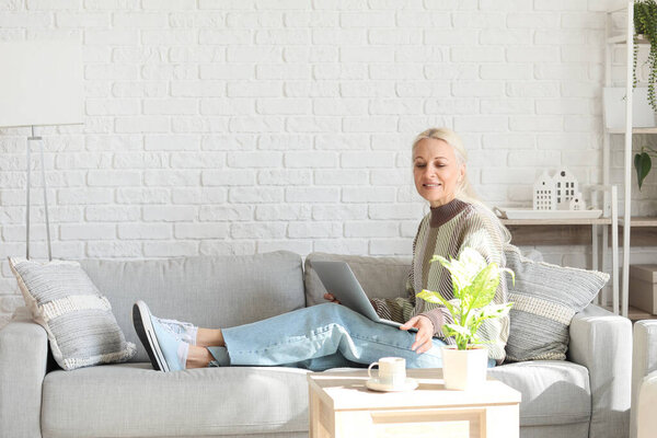 Mature female programmer with laptop taking cup of coffee from table at home