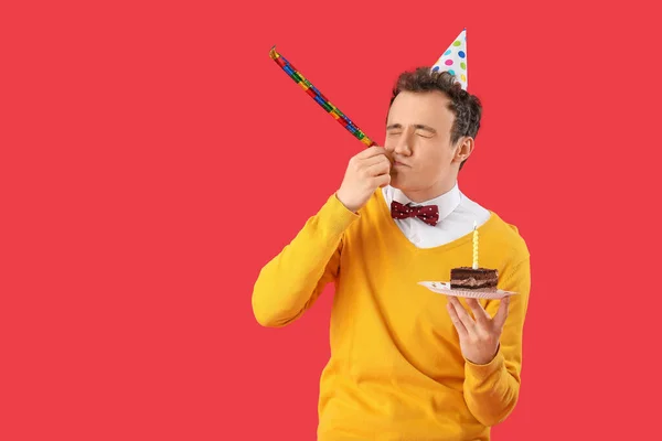 stock image Young man with birthday cake and party whistle on red background