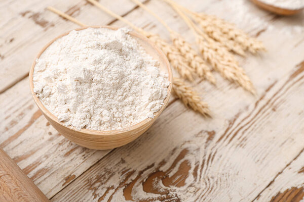 Bowl with wheat flour and spikelets on white wooden table, closeup