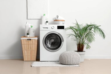 Çamaşır makinesi, deterjan şişeleri, çamaşır sepeti ve beyaz duvarın yanında puf.