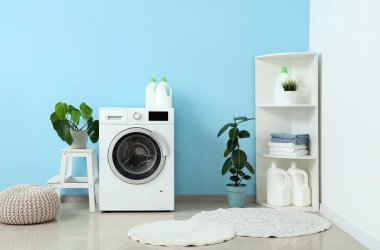 Çamaşır makinesi, raf ünitesi, ev bitkileri ve mavi duvarın yanındaki puf.
