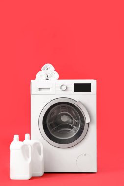 Çamaşır makinesi, deterjan şişeleri ve kırmızı arka planda rulo havlular.