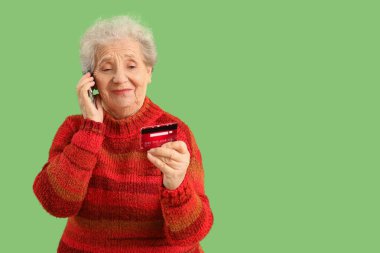 Kredi kartı olan yaşlı bir kadın yeşil arka planda cep telefonuyla konuşuyor.