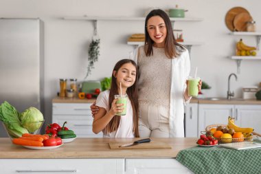 Küçük kız hamile annesiyle mutfakta yeşil smoothie içiyor.