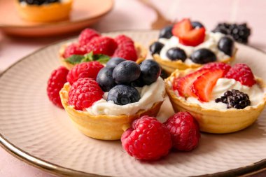 Plate of tasty tartlets with whipped cream and berries on pink background, closeup