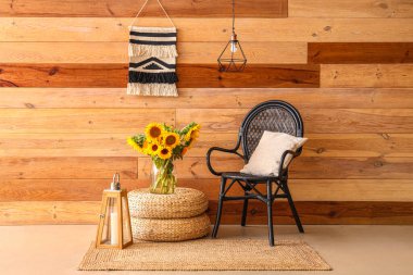 Vase with beautiful sunflowers on wicker pouf and armchair near wooden wall
