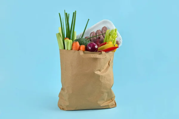 Paper bag full of fresh vegetables and sausages on blue background