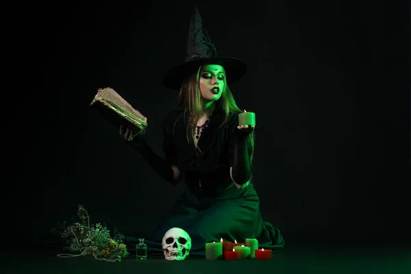 暗い背景に呪文の本とろうそくが付いている美しい若い魔女 — ストック写真